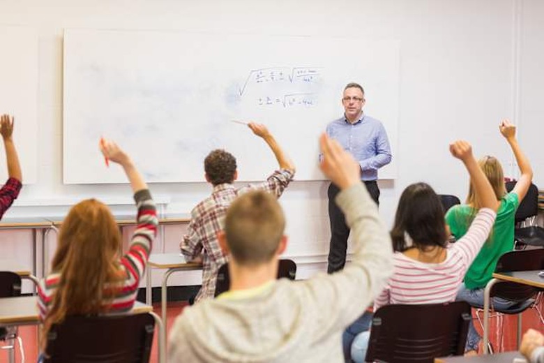 Rear view of students with hands raised with a teacher in the classroom