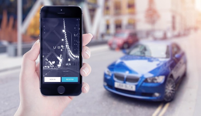 Uber application startup page on the Apple iPhone 5s display in female hand. Blurred street view with car and flare sun light on