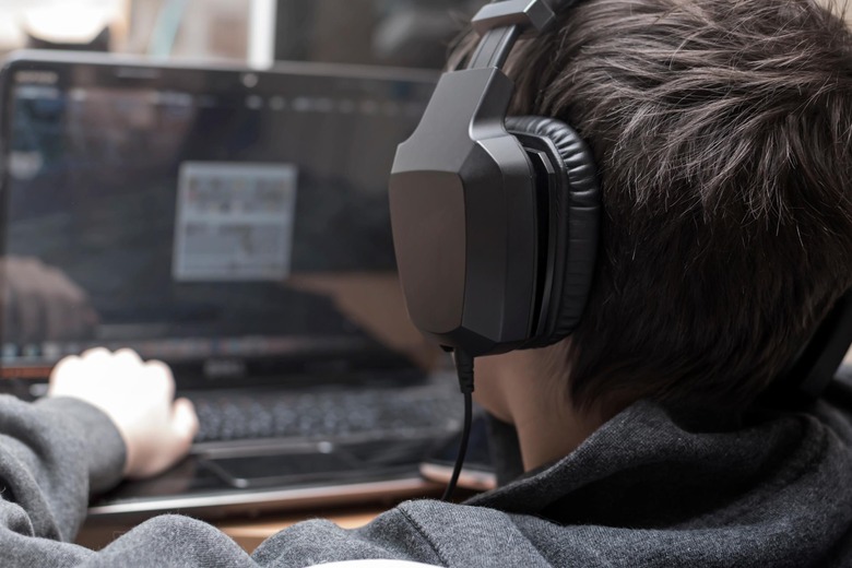 Boy with headphones is viewing the monitor of computer during playing computer games, chatting on the internat, watching a video or reading, side view, the face is not visible