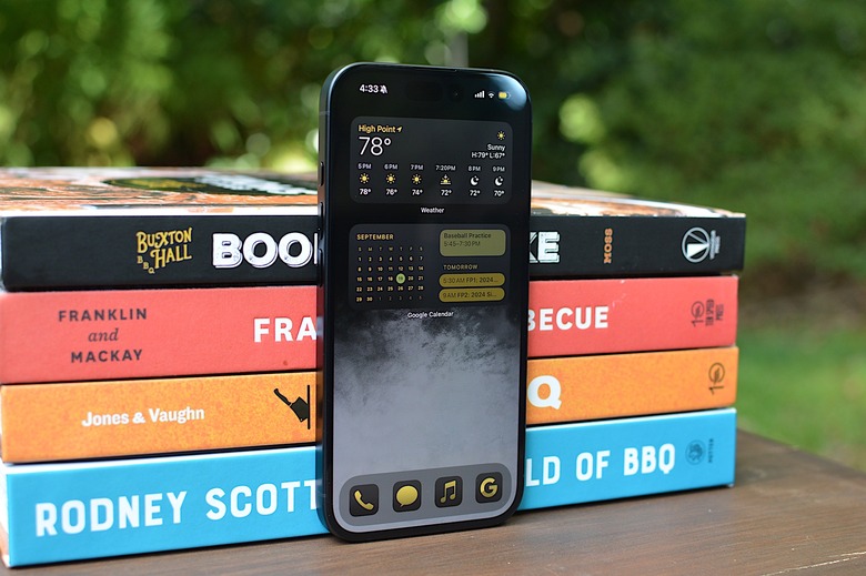 An iPhone running iOS 18 leaning against a stack of books outside.