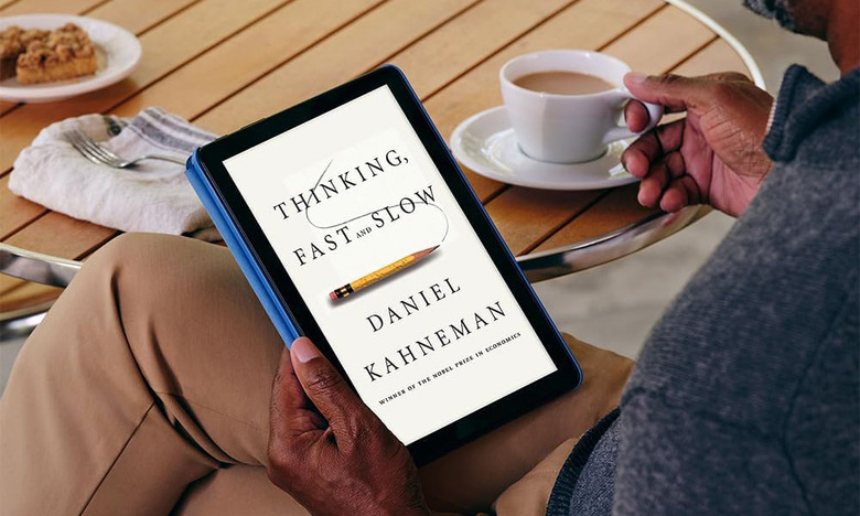 A person holds the Amazon Fire HD 10 tablet that displays the cover of an ebook. The person is drinking a latte, or maybe just coffee with cream. 