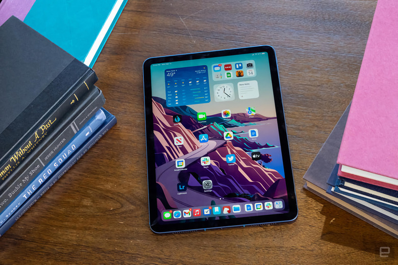Photo of the iPad Air sitting on a wooden desk or table next to piles of books. Home screen visible on the iPad.