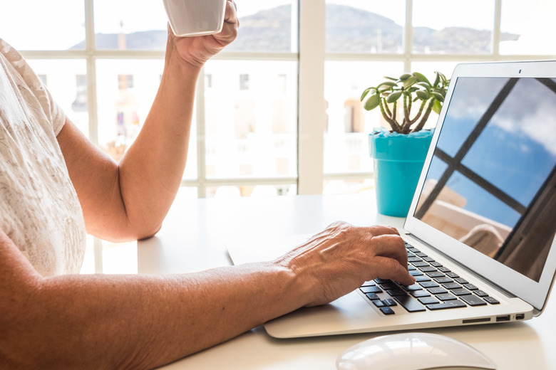 Close-up view of woman's hand typing on laptop keyboard while holding a cup of tea or coffee. Nomadic mature female enjoying tech and social working home