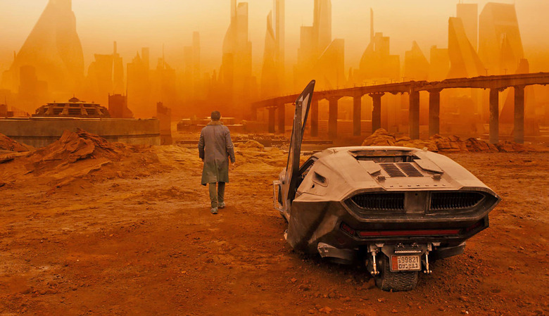 A still from Blade Runner 2049 showing Ryan Gosling walking away from a futuristic vehicle in a ruined urban landscape filled with thick, orange smog