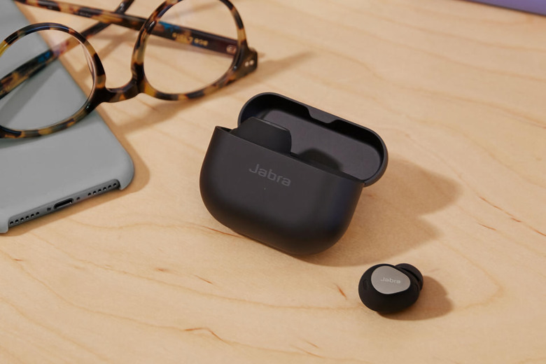 Black wireless earbuds sitting on a desk. One bud is out of the case, sitting on its own, while the case (with the other bud) sits next to it. Glasses and a smartphone are nearby.