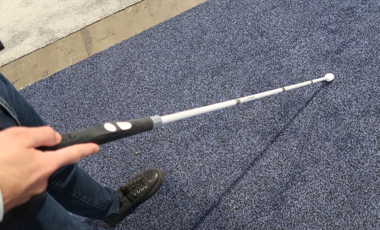 The WeWalk Smart Cane 2 is shown from the angle of the person holding it. Their hand can be seen gripping the handle and the cane is held out in front over a navy blue carpet