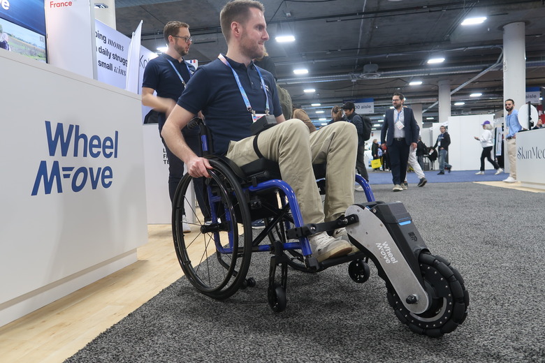 A man sits in a manual wheelchair with an add-on attached that gives it a large central front wheel and raises the casters off the ground