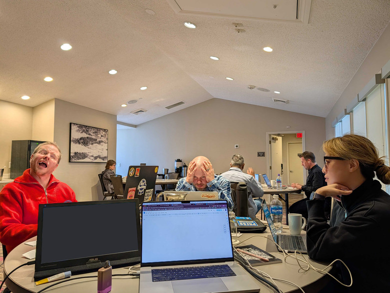 Three people with strange expressions sit around a table with laptops in front of them.