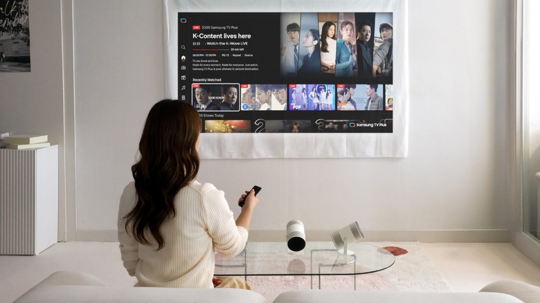 A woman is pictured from behind sitting on a white sofa with a remote pointed to a streaming screen projected on a white sheet, with the Freestyle+ projector in front of her on a glass coffee table