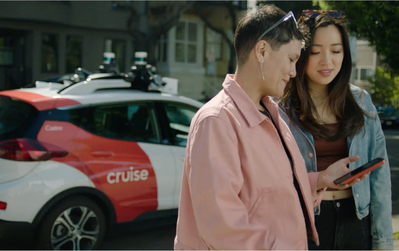 Two persons in the foreground, both looking down on a smartphone. In the background is a Cruise robotaxi painted red and white.