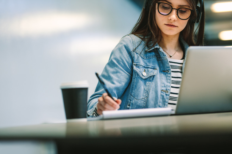 University student writing in a book while sitting at desk with laptop and coffee up at college campus. Female student studying at college library.