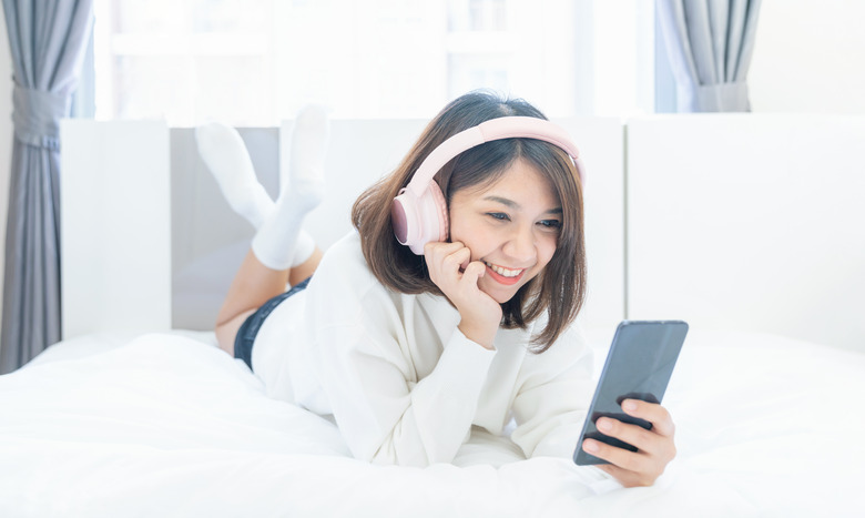 Beautiful young woman with headphones relaxing on the bed, Female listening to music using smart phone, Chill out and leisure concept