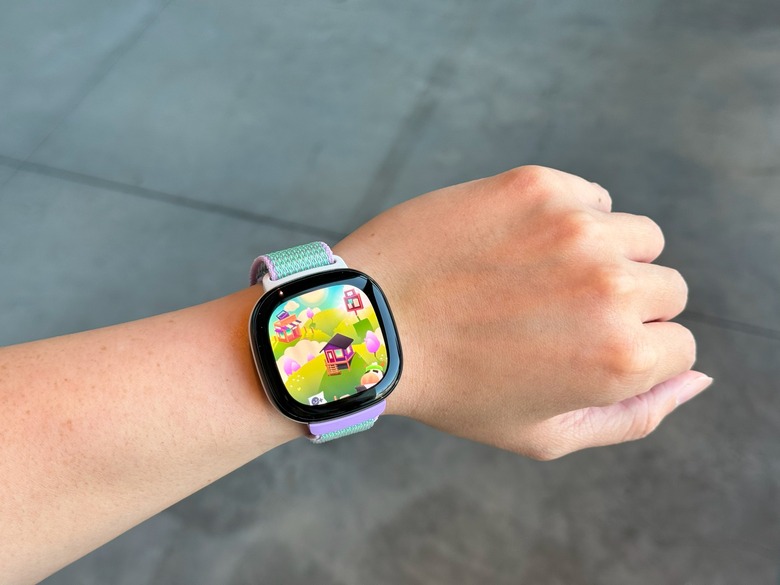 The Fitbit Ace LTE on a wrist held in mid-air, with a house sitting in a picturesque valley on its screen.