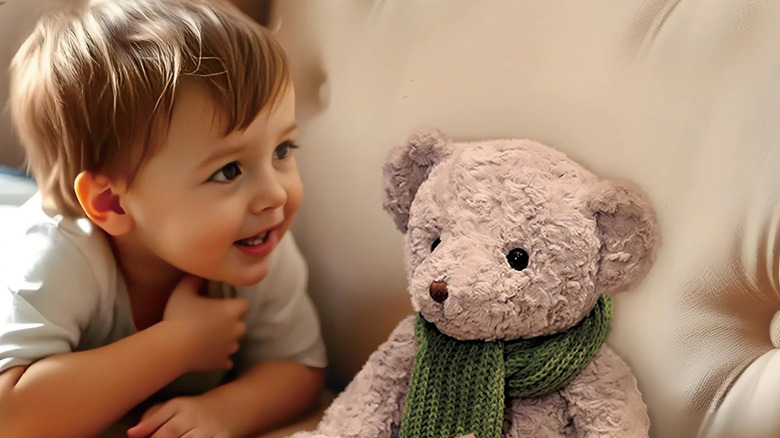 Marketing photo of a child grinning, looking at a teddy bear on a counch.