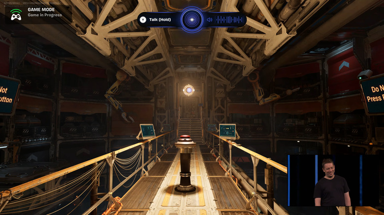 Still from an onstage Epic demo. A dimly lit catwalk with a red button in the middle. Presenter camera feed in a box on the lower right.