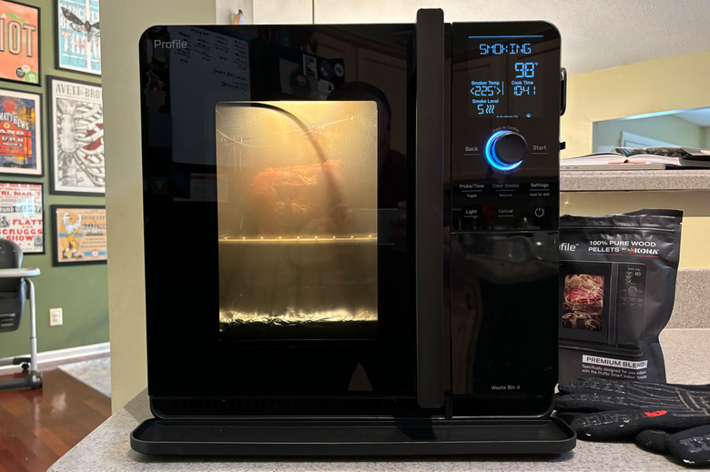 GE Profile Smart Indoor Smoker from the front with the interior light on, sitting on a kitchen counter. A pork butt is being smoked inside.