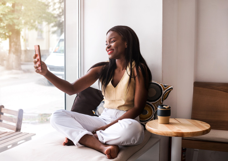 Attractive african girl makes selfie at the windowsill at cafe with reusable cup of coffee.