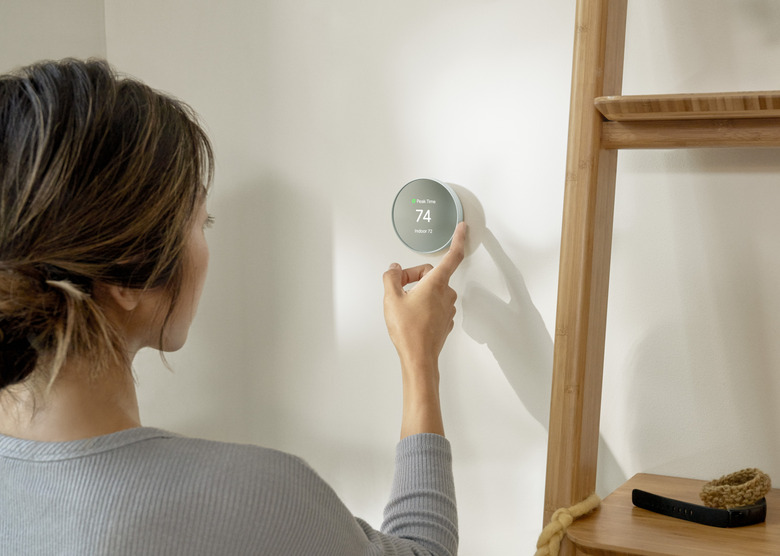 A person using their right index finger to adjust the temperature on a Nest Thermostat, which shows the temperature at 74 degrees on its screen. It also displays a green leaf symbol above the numbers, with small white text next to it.