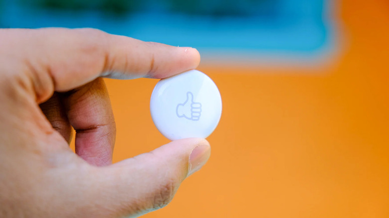 A person’s left hand holding an Apple AirTag (white disc shape) with a thumbs-up emoji inscription on it. Orange and blue blurred background.