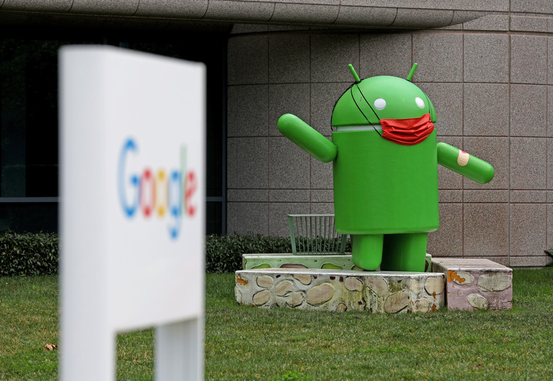 An image from outside of the Google campus building in Mountain View, CA. showing an out of focus Google sign in the foreground and an Android statue with an ad-hoc face mask behind it.

