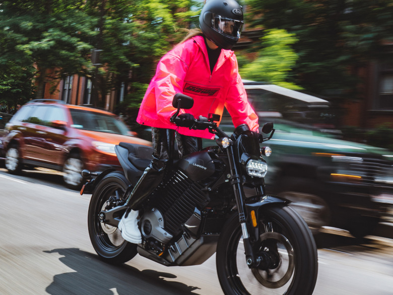 A person in a flamingo-pink windbreaker riding the LiveWire S2 Del Mar electric motorcycle down a city street. Cars are parked behind in front of trees and visible sections of city buildings.
