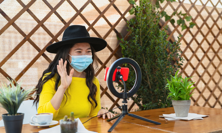 Happy Asian woman wearing face mask and vlogging online using smartphone cam and influencer led while drinking a coffee sitting in bar during corona virus outbreak