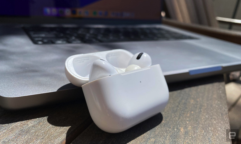 A pair of AirPods in the case sit on a wooden table next to an open MacBook. 