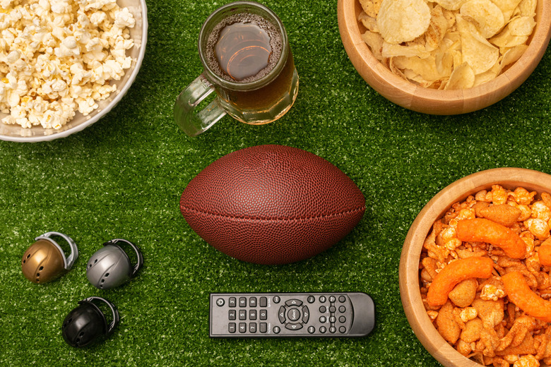 Super bowl flat lay photography with snacks, beer, american football ball, little helmets and remote control on green grass background