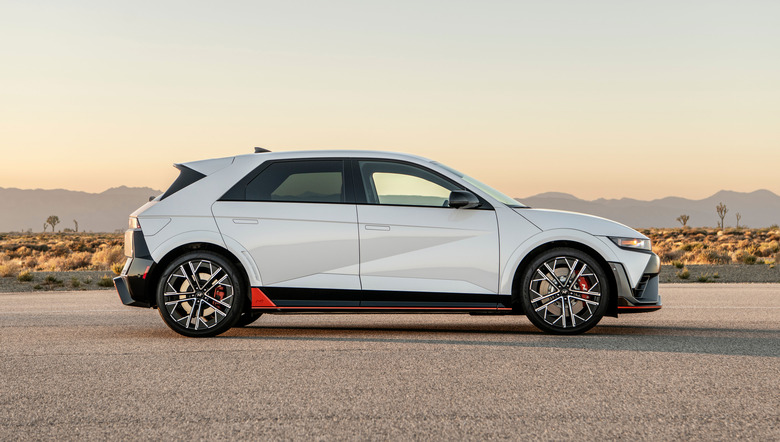 A white Hyundai Ioniq 5 seen in profile is parked in the middle of a paved lot in a desert area with a hazy dawn or sunset and low hills in the distant background. 