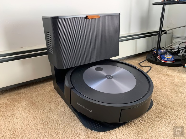 Photo of the iRobot Roomba j7+ robot vacuum sitting against a wall (with heat vent directly behind it) on a carpeted floor. It’s docked in its cleaning station.