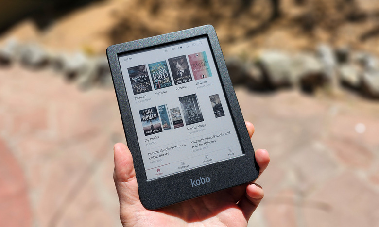 A person holds the Kobo Clara Colour in the sun. The color covers of some books are displayed on the screen. There are rocks in the background. 