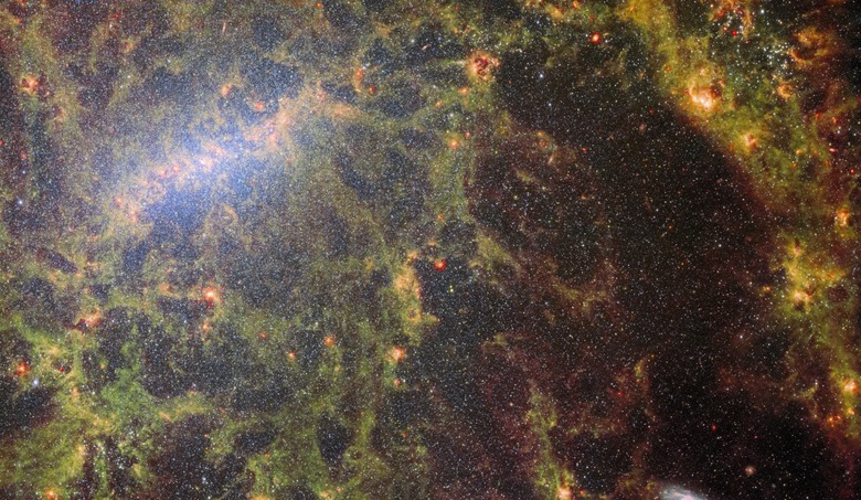 A close-in image of a spiral galaxy, showing its core and part of a spiral arm. Thousands upon thousands of tiny stars that make it up can be seen, most dense in a whitish bar that forms its core. Clumps and filaments of dust form an almost skeletal structure that follows the twist of the galaxy and its spiral arm. Large, glowing bubbles of red gas are hidden in the dust.