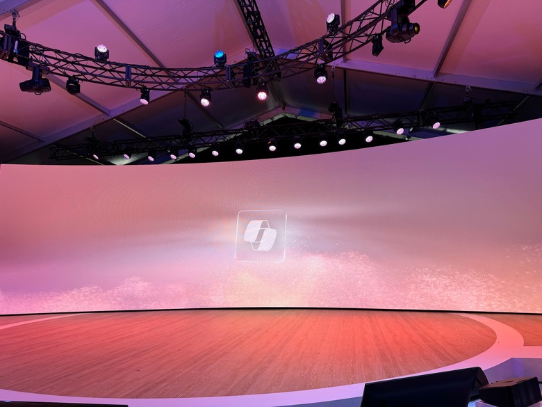 A round stage with wooden floors and a curved screen in the back is showing rosy clouds and the Copilot logo overlaid.