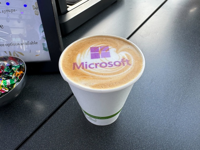 A cup of coffee with milk art on top and a purple Windows logo with the word Microsoft also on top of the milk art.