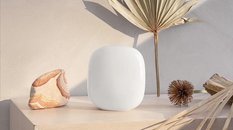 The Google Nest WiFi Pro - 6E is smooth and white and sits on a white oak countertop with a rock and other natural-looking tchotchkes nearby.  