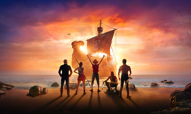 Promotional art for Netflix’s live-action ‘One Piece.’ Five crew members face a shoreline with their backs to us. In the center, Monkey D. Luffy (Iñaki Godoy) stands with his arms raised triumphantly. A pirate ship approaches on the seas in the background.