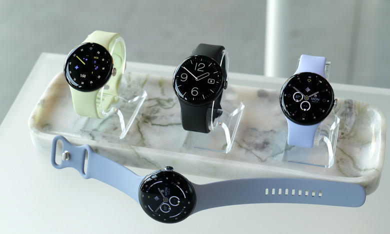 A trio of Pixel Watch 4 devices each on their own stand, laid out on a table, with a fourth Pixel Watch 4 laying horizontally in front of them.