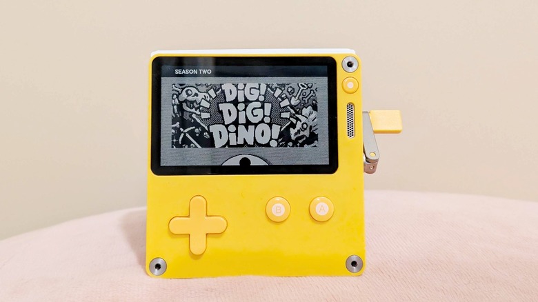 A yellow Playdate console pictured standing on a soft pink surface displaying the title card for the game Dig! Dig! Dino!