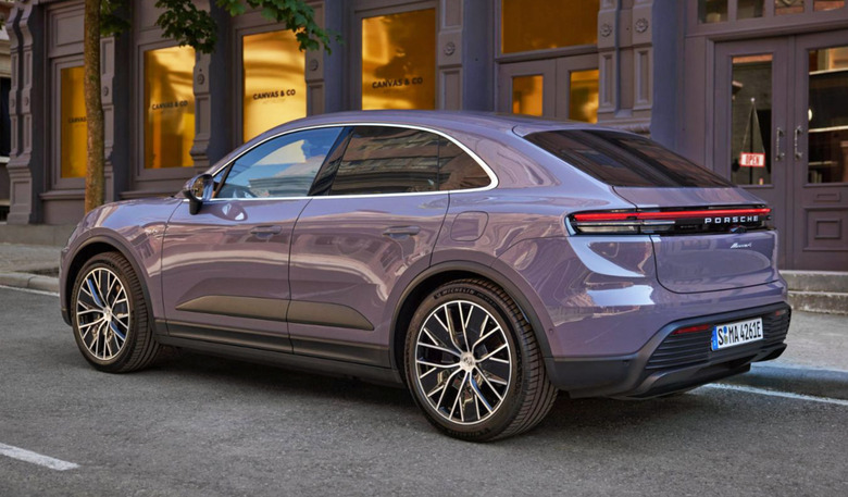 A purplish-grey Porsche’s all-electric Macan SUV is parked on the side of a road in front of doorways and windows.