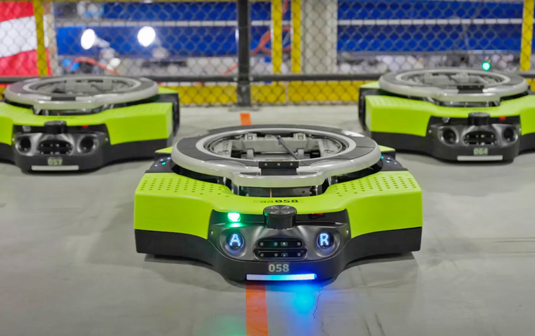 A trio of Amazon's fully autonomous Proteus warehouse robots face forward towards the camera as they sit on the floor, looking like a yellow Roomba vacuum.
