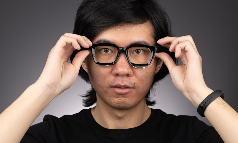Cornell University researcher Ruidong Zhang modeling a prototype glasses add-on, which includes microphones and speakers below the lenses. He is looking straight into the camera while holding the arms of his worn glasses.