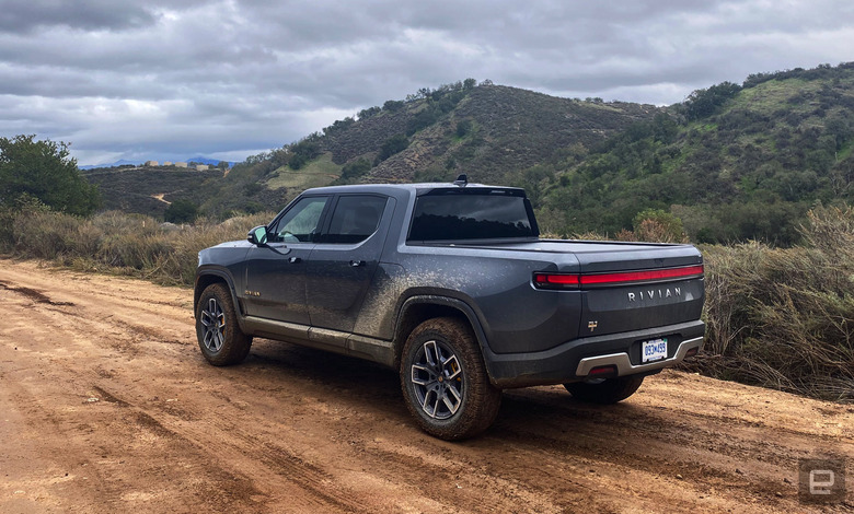 A dark grey Rivian R1T drives up a dirt road that's headed upwards with hills in the background.