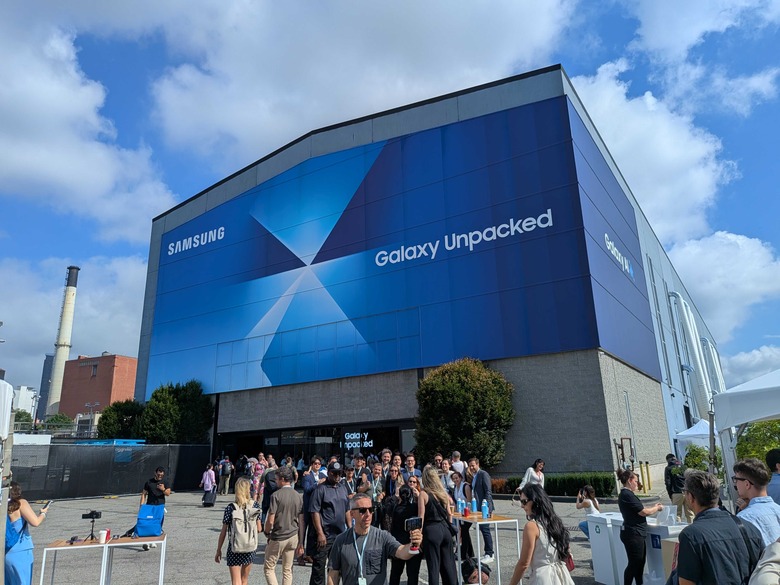 A line of people standing in front of a large building with a banner on it showing the Samsung Galaxy Unpacked invitation image.