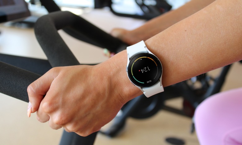 Closeup of a person's wrist, wearing a Samsung Galaxy Watch and riding on a Peloton exercise bike in a gym. A heart rate of 124 bpm is displayed on the watch.