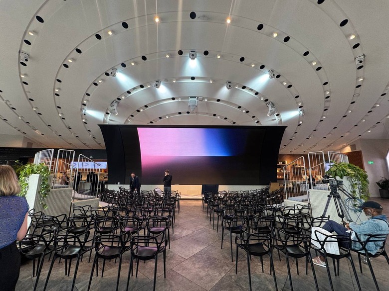 An event space with a screen in front and two sets of empty chairs. A camera tripod is at the bottom right of the image.