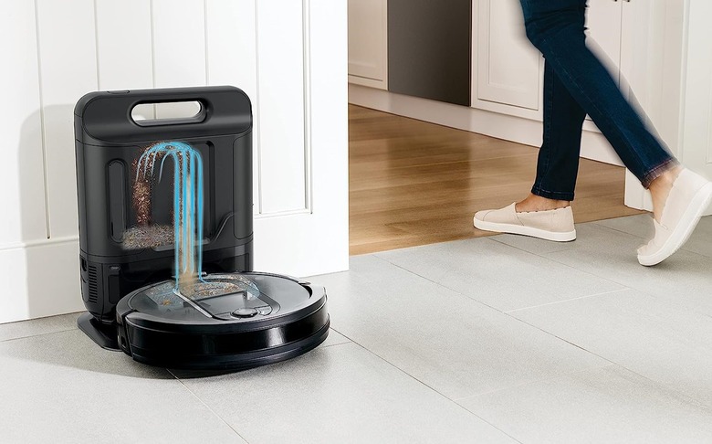 A robot vacuum docked in its base, with an illustration showing the process of the vacuum emptying itself. A person walks by in the background.