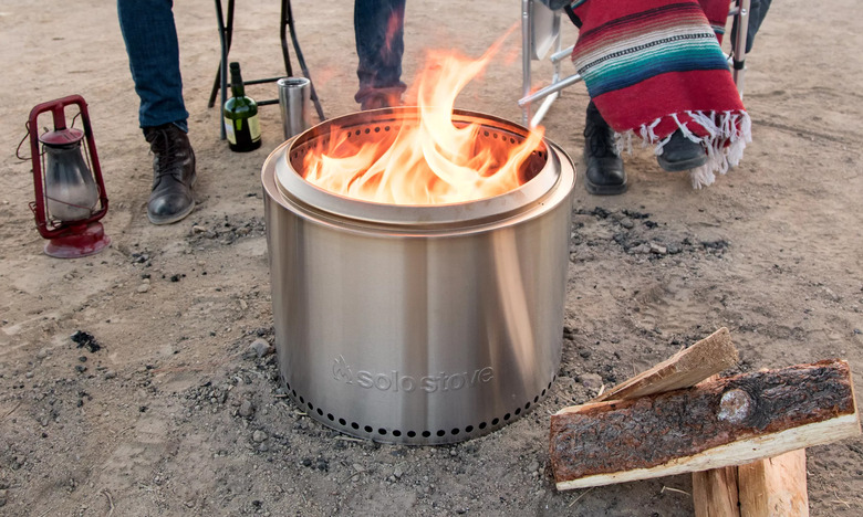 The Solo Stove Bonfire burns wood on a sandy ground. Two people sit in folding chairs in the background. One covers their legs with a colorful blanket. 