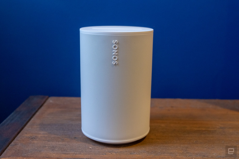 Photo of the Sonos Era 100 smart speaker (white) sitting on a wooden table in front of a purple wall.