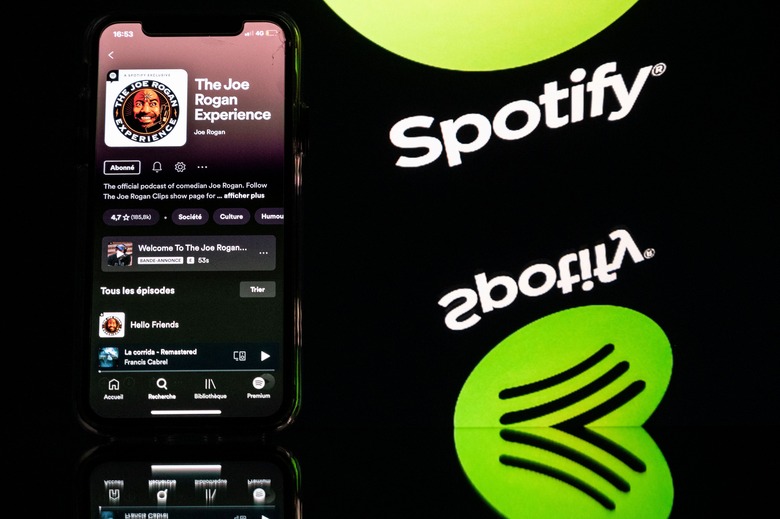 This picture taken in Toulouse, southwestern France, on January 31, 2022 shows a smartphone displaying the Joe Rogan podcast and a screen displaying the Spotify's logo. - Caught in a disinformation maelstrom, Spotify announces it will guide listeners of Covid-19 podcasts to established facts about the pandemic. This comes after artists pulled their songs in protest at the platform given on Spotify to podcaster Joe Rogan, accused of spreading damaging myths about the pandemic. (Photo by Lionel BONAVENTURE / AFP) (Photo by LIONEL BONAVENTURE/AFP via Getty Images)