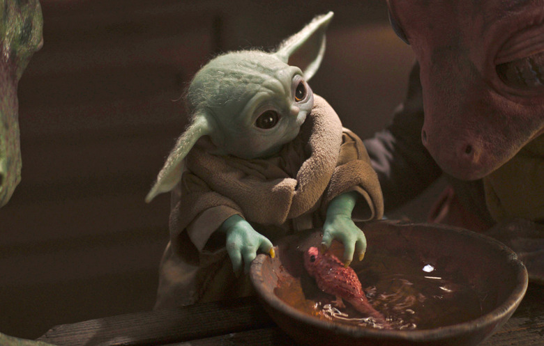 Grogu plays with a small pink fish-like creature in a bowl of water as a larger creature looks on.